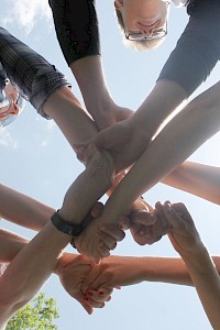 A group of people standing in a circle with their hands joined in the center, against a blue sky backdrop.