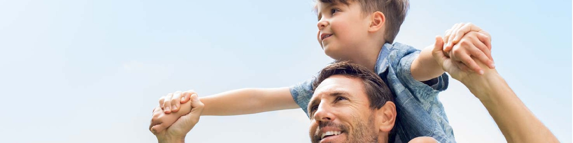 A man is carrying a child on his shoulders against a blue sky, holding hands and smiling.