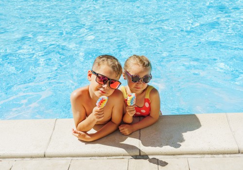 Two children in sunglasses enjoying colorful popsicles by a pool edge under a bright sunny day, wearing swimwear.