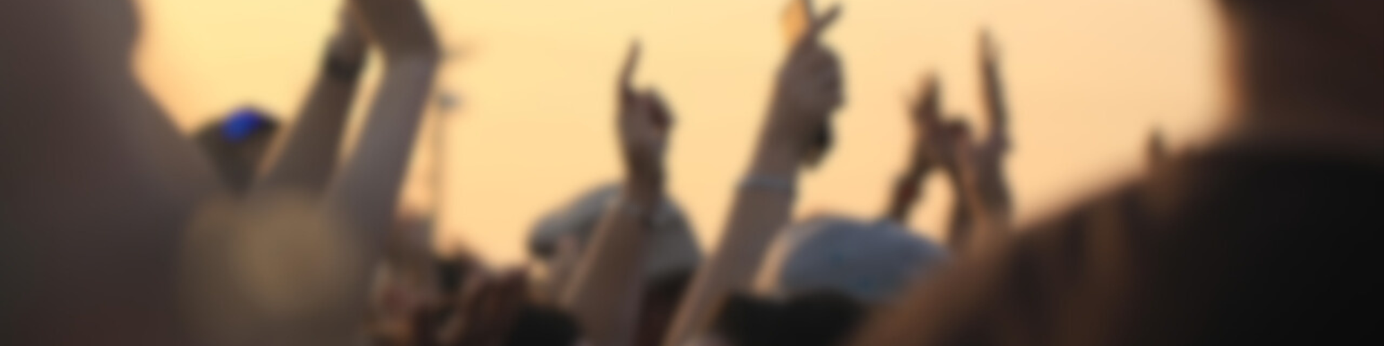 Blurry image of a crowd outdoors, hands raised, likely at a concert or event during sunset.