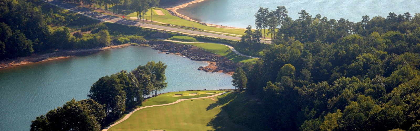 An aerial view of a scenic golf course with lush greenery, water features, and surrounded by a large body of water and trees.