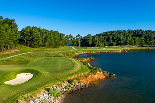 A scenic golf course with a green, sand trap, and surrounding water and trees under a clear blue sky.