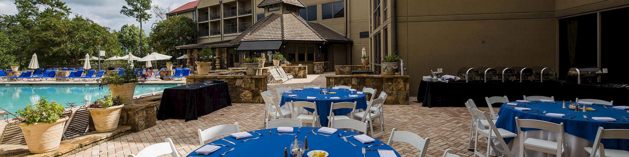 An outdoor dining setup by a pool with round tables covered in blue tablecloths and white chairs, located near a multi-story building.