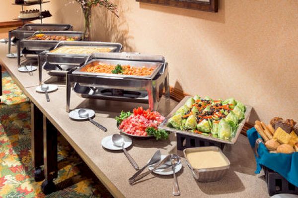 A buffet setup featuring chafing dishes with various hot foods, a tray with fresh salad, cut vegetables, and a basket of bread and muffins.