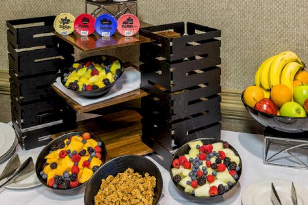 A table with bowls of fruit salads, granola, and a variety of whole fruits, alongside yogurts on a wooden display stand, and plates and cutlery nearby.