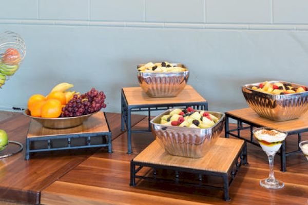 The image shows a buffet table with a variety of fresh fruits in bowls, including grapes, oranges, and mixed fruit salads, along with parfaits in glasses.