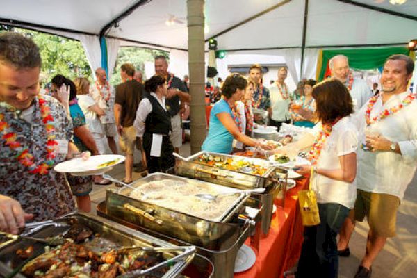 A group of people are gathered at a buffet line in a tented outdoor setting, enjoying a vibrant gathering with food and drinks, wearing casual attire.