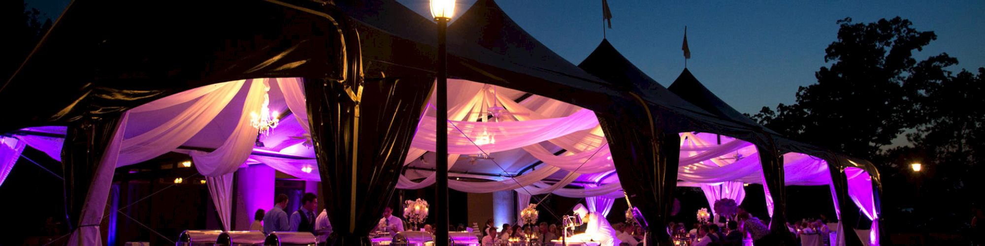 An outdoor event under a tent with white drapes and purple lighting, featuring tables set up and guests mingling under a night sky ending the sentence.