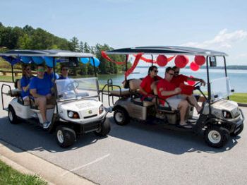 Two decorated golf carts, each carrying four people, drive along a lakeside road in a festive setting, with blue and red decorations.