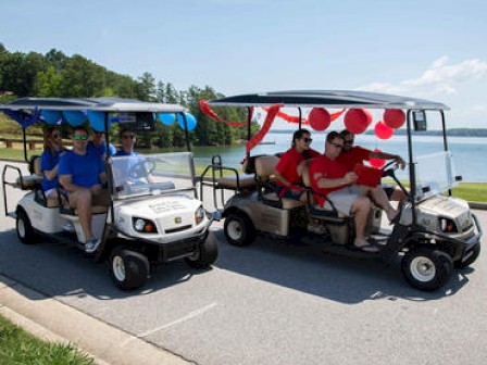 Two decorated golf carts, each carrying four people, drive along a lakeside road in a festive setting, with blue and red decorations.