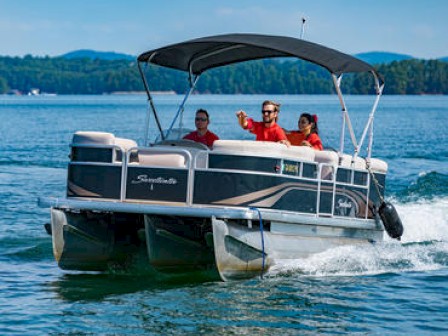 Three people are on a pontoon boat, enjoying a day on the water. The boat is moving, and they are under a canopy on a sunny day with a scenic backdrop.