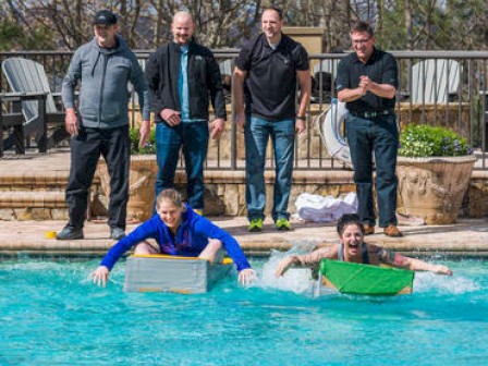 A group of people watch from behind a pool fence while two individuals race in the water using makeshift cardboard boats, displaying enthusiasm and fun.