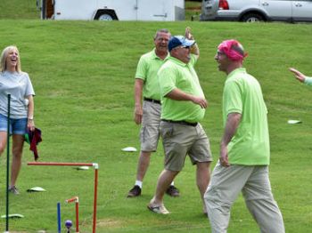 Group of people enjoying an outdoor activity on a grassy field; some wear matching green shirts, and one wears a pink bandana.