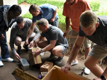 A group of individuals gathers around, examining and handling wooden boxes and containers, possibly engaging in an outdoor activity or project.