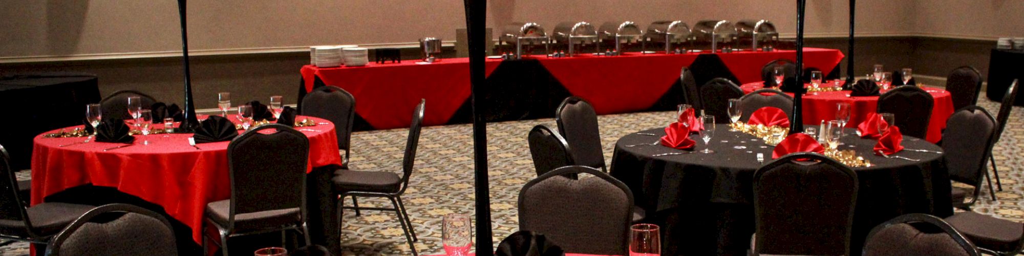 A banquet hall with round tables adorned with red tablecloths, tall red floral centerpieces, and folded black napkins, set for an event.