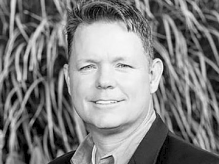 A grayscale portrait of a smiling man, wearing a blazer and collared shirt, with foliage in the background.