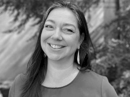 A black and white photo of a smiling person with long hair, wearing earrings and a top, standing in front of a blurred background with trees.