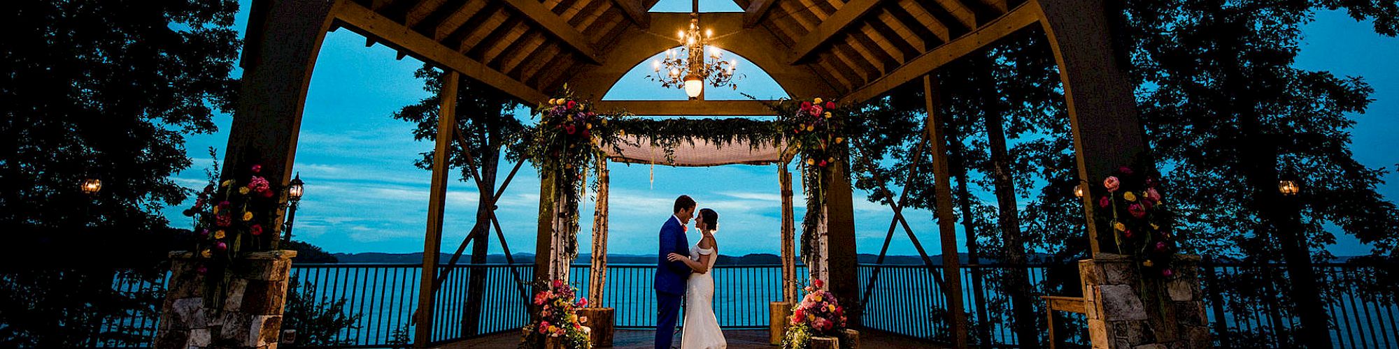 A couple shares a dance under a beautifully lit wooden gazebo with floral decorations, surrounded by trees and overlooking a body of water.