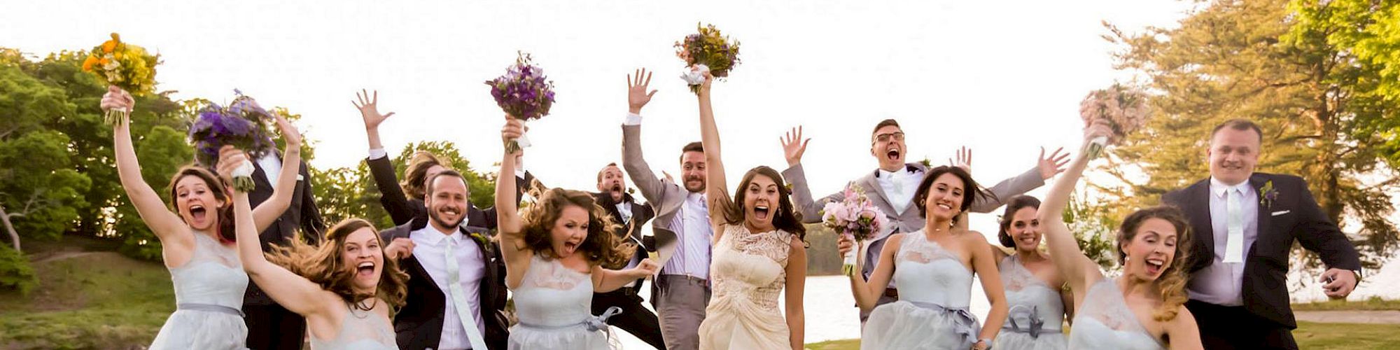 A wedding party is captured mid-jump. The bride, groom, bridesmaids, and groomsmen are joyously celebrating in an outdoor setting, dressed formally.