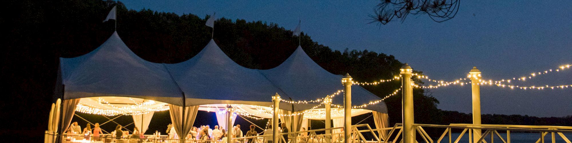 This image shows an evening event under a white tent by the water, adorned with string lights, creating a festive atmosphere.