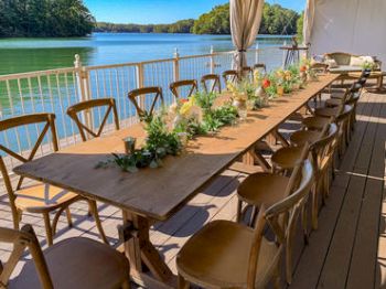An outdoor table set for an event with wooden chairs and floral arrangements overlooks a serene body of water with greenery in the background.