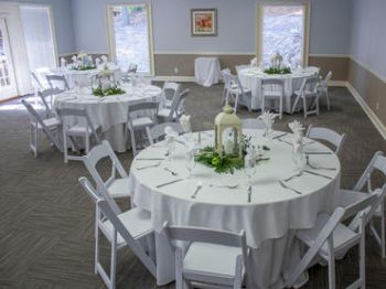A decorated room with round tables set with white tablecloths and chairs, prepared for an event or gathering, featuring centerpieces and place settings.