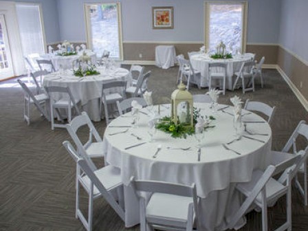 A decorated room with round tables set with white tablecloths and chairs, prepared for an event or gathering, featuring centerpieces and place settings.