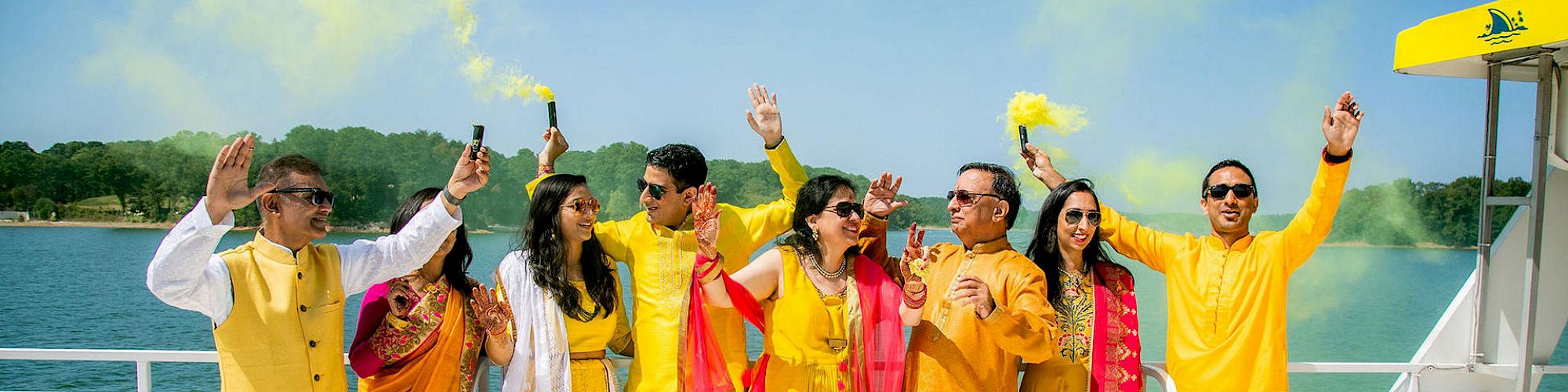 A group of people dressed in vibrant yellow and orange outfits, holding colored smoke sticks, standing on a boat with water and trees in the background.