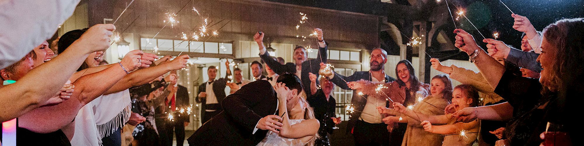 A couple sharing a kiss in the center, surrounded by people holding sparklers, likely celebrating a wedding or similar event, at night.