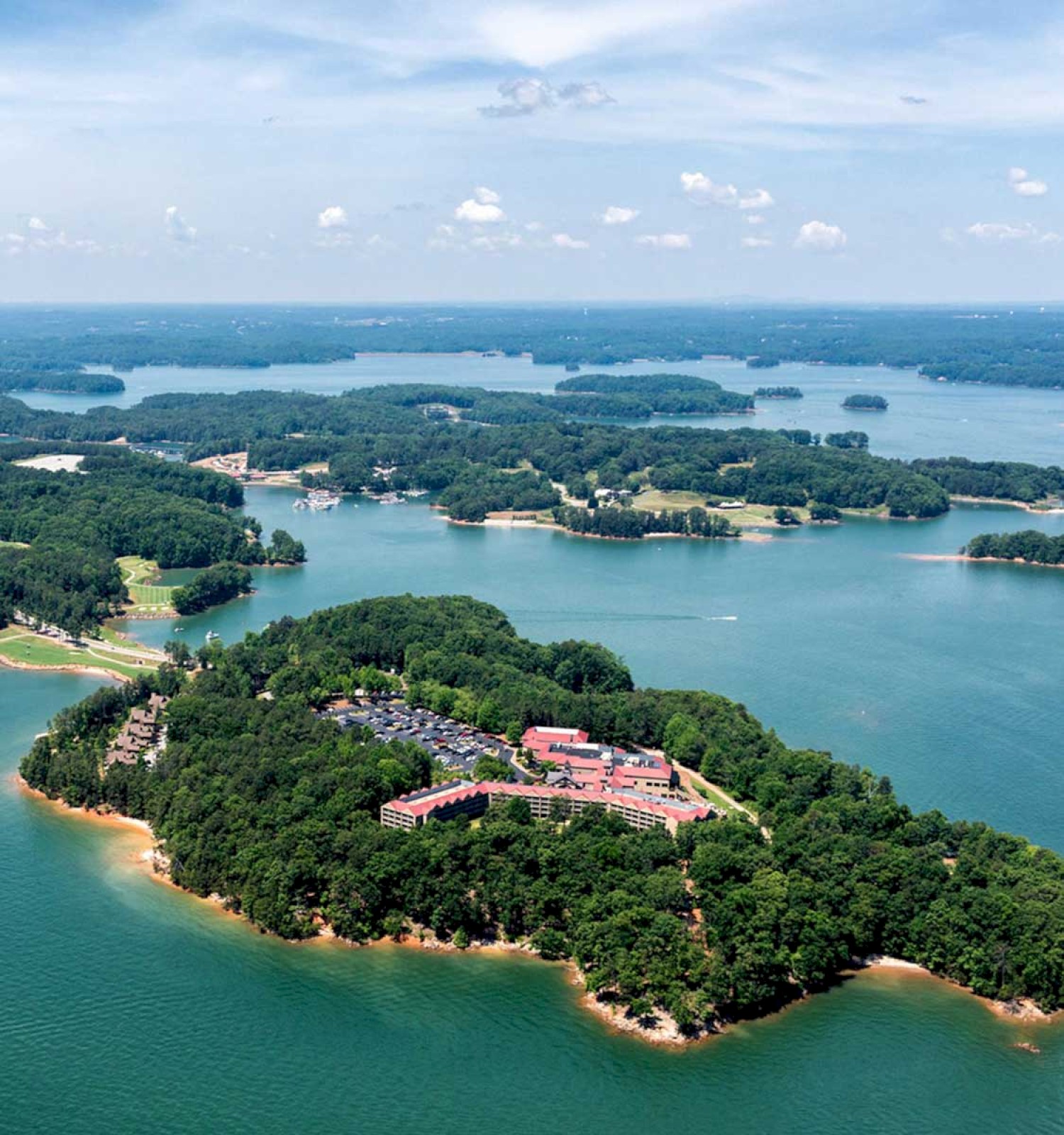 Aerial view of a lush, green island in a lake with a complex of buildings and parking, surrounded by water and distant forests.