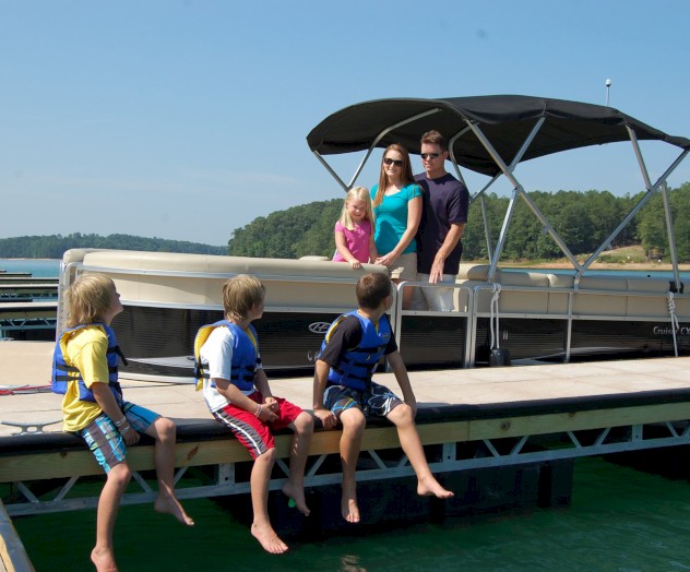 A family is enjoying time by a boat on a dock, with three children sitting at the edge and two adults standing nearby on the boat.