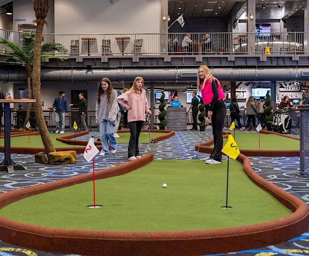 Three people are playing mini-golf indoors, surrounded by others in a lively environment with colorful carpet and artificial turf.