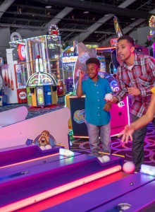 People enjoying arcade games inside a well-lit gaming center, with various machines in the background.