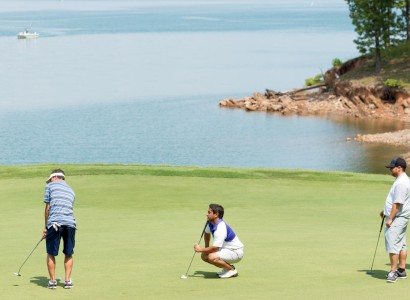 Four people are playing golf near a body of water, with a boat in the background.