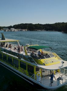 A green and yellow boat with multiple passengers navigates a lake surrounded by forested islands and a distant waterfront area.
