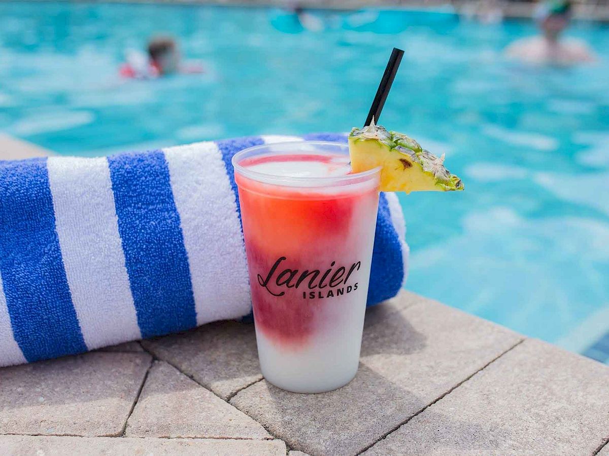 A colorful drink with a pineapple garnish and a straw sits by a poolside on sunlit tiles next to a blue and white striped towel.