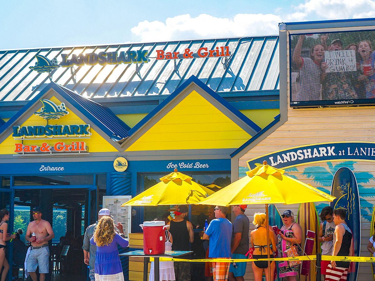 This image shows the entrance of LandShark Bar & Grill with people lined up outside under yellow umbrellas, featuring surfboards and a large screen above.