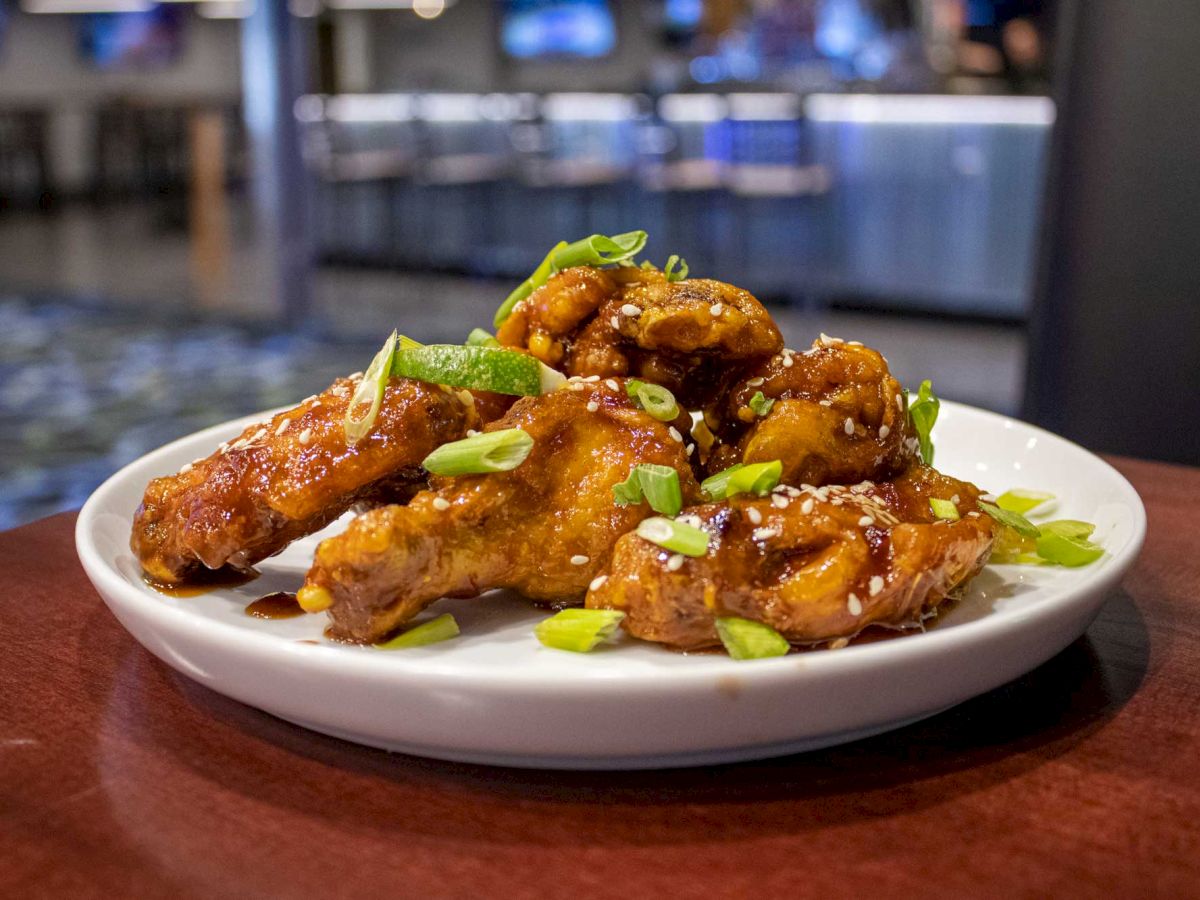A plate of sauced chicken wings garnished with sesame seeds and chopped green onions, set on a wooden table in a restaurant setting.