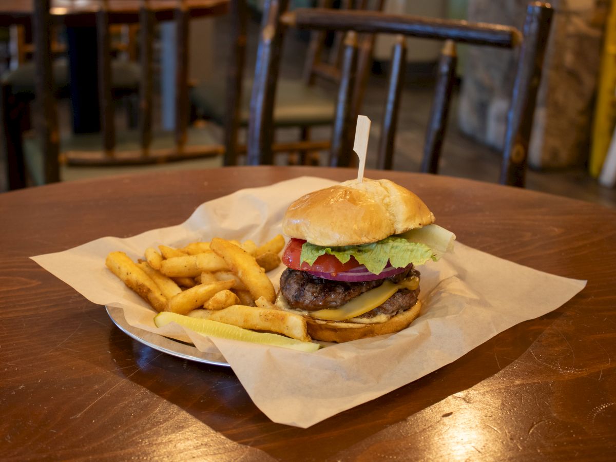 A cheeseburger with lettuce, tomato, and onion sits on a plate with fries on a wooden table. The table has rustic chairs around it.