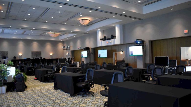 An empty conference room with rows of tables and chairs, television screens, lighting equipment, and a patterned carpet.