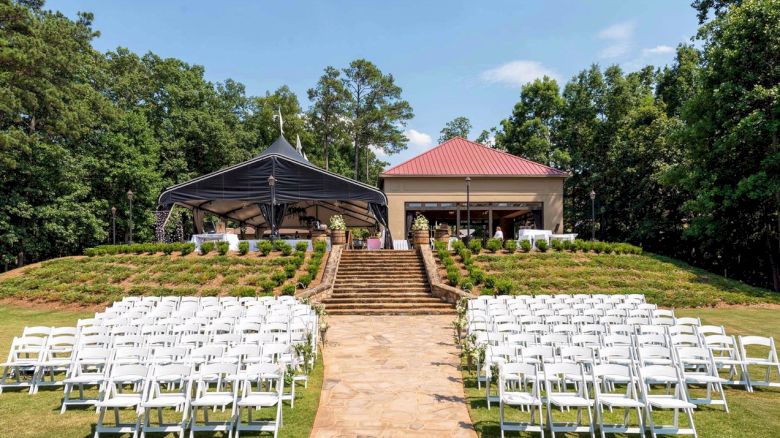 An outdoor wedding setup with rows of white chairs facing a staircase leading to a small building and a shaded area, surrounded by greenery.