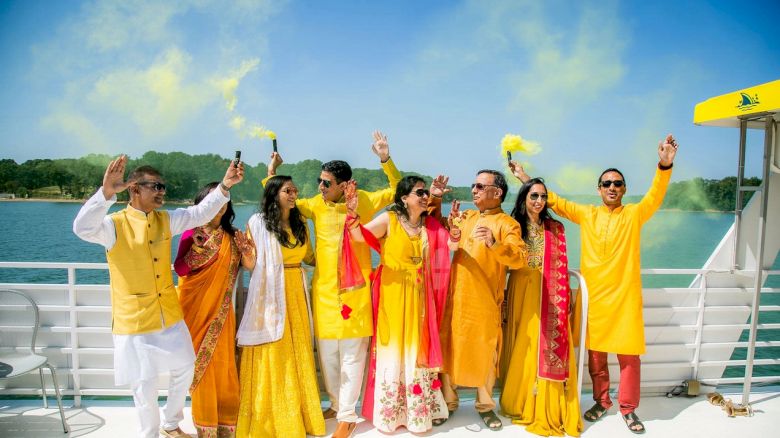 A group of people dressed in colorful traditional attire celebrate with yellow smoke on a boat by the water under a sunny sky.