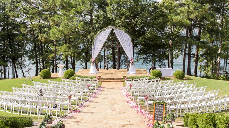 An outdoor wedding setup by a lakeside, featuring rows of white chairs, a decorated arch, and a stone path lined with flower petals, in a forested area.