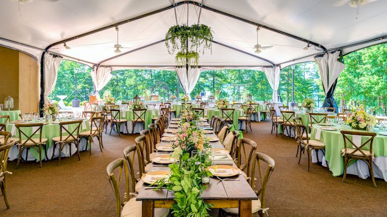 The image shows a beautifully decorated event tent with elegant tables and chairs, featuring a long centerpiece and greenery, set against a backdrop of trees.