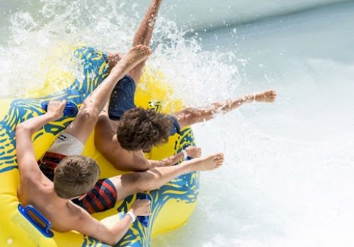 Three boys are enjoying a water ride on a yellow inflatable raft, splashing through a wave in a water park setting.