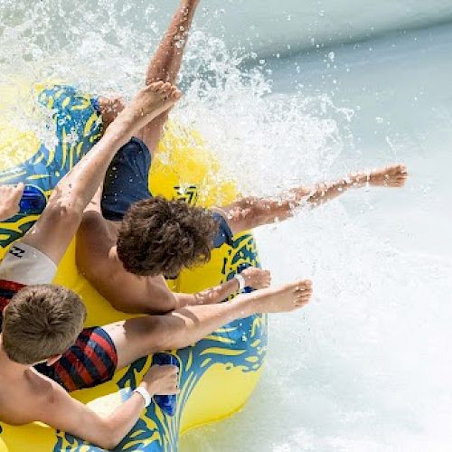 Three boys are enjoying a water ride on a yellow inflatable raft, splashing through a wave in a water park setting.