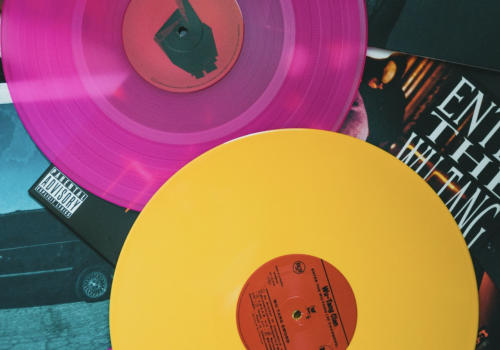 Two colored vinyl records, one pink and one yellow, lie atop album covers, featuring various designs and text.