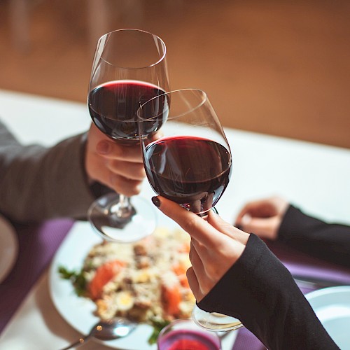 Two people clink glasses of red wine over a table setting with plates and a dish in the background.