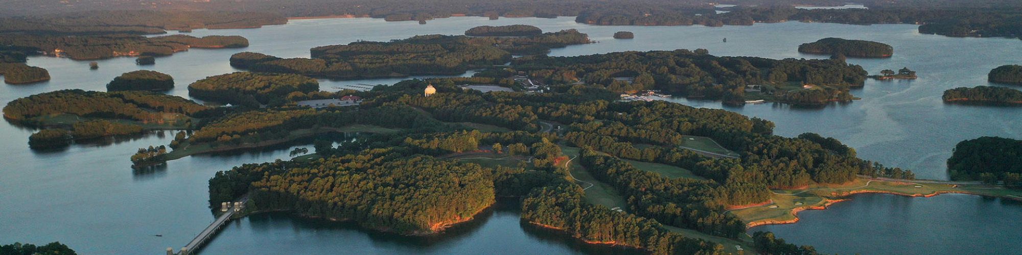 Aerial view of a lake with numerous small, forested islands at sunset. The water is calm and the surrounding landscape is lush and green.