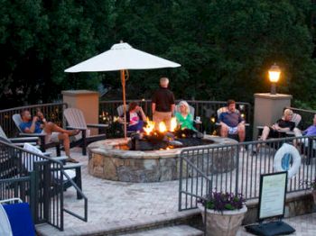 A group of people are sitting around a fire pit on a patio, surrounded by trees, with an umbrella and lit lamps creating a cozy ambiance.